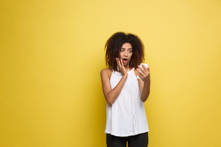 Shocked woman wearing ear phones looking at her smartphone against a yellow wall